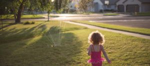 Little girl running through front yard to a sprinkler.