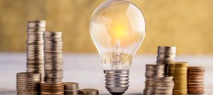 Lit lightbulb surrounded by stacks of coins at different heights.